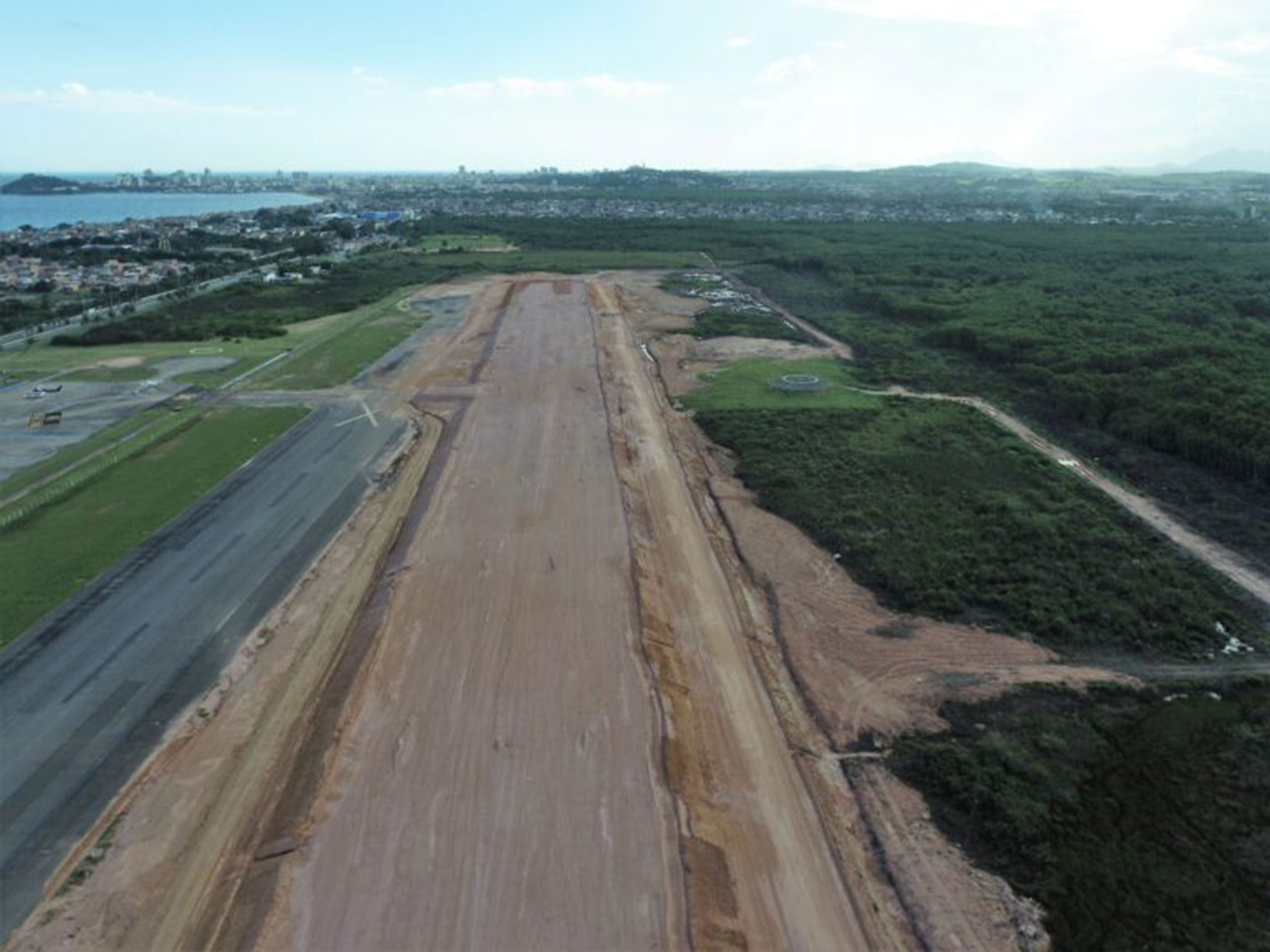 Photo of a construction site on the new runway at Macaé Airport in Brazil from 2024. Various construction equipment and machines are active on the runway. Part of the airport building can be seen in the background. Engineers in high-visibility vests monitor the construction progress.